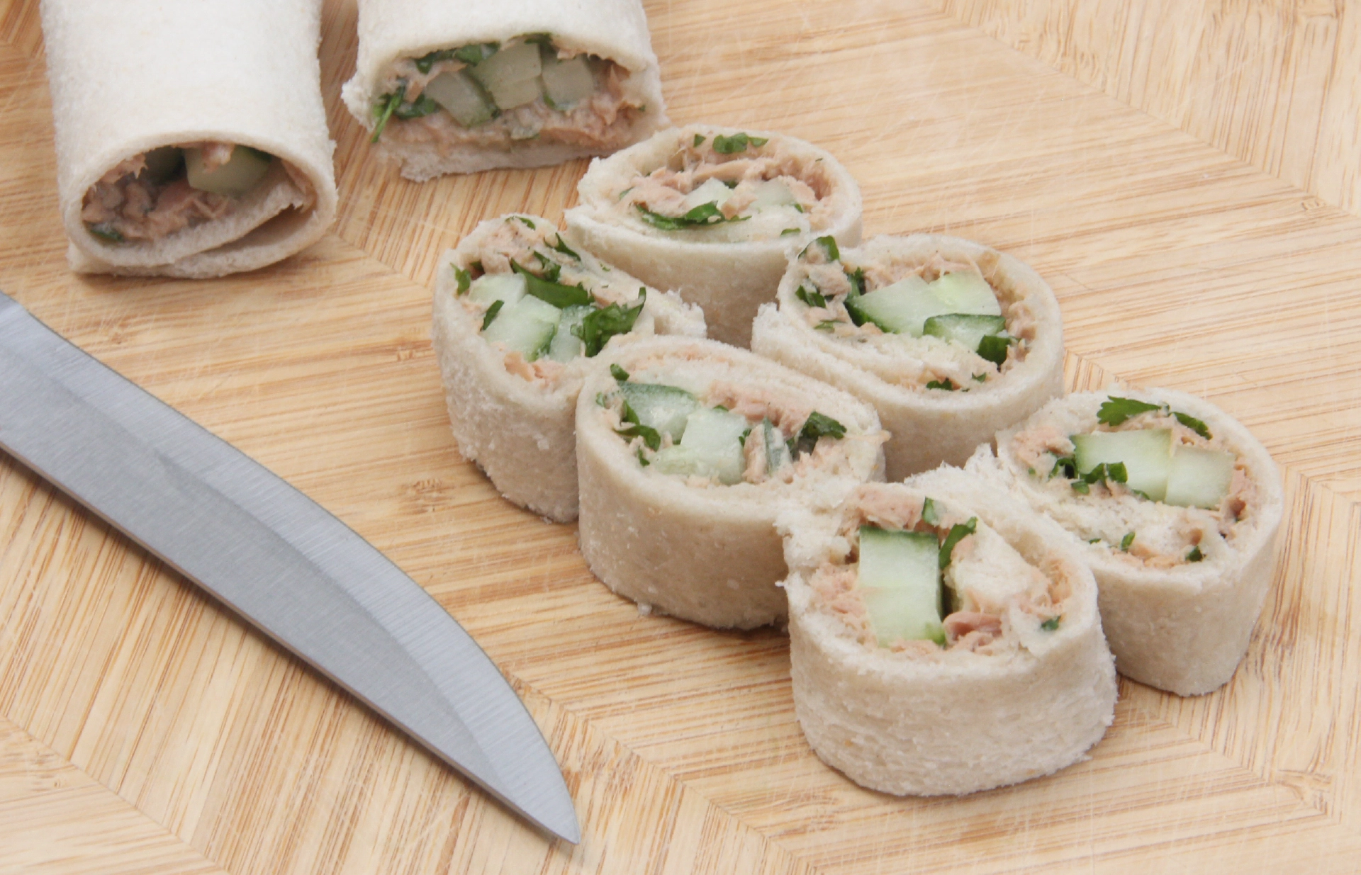 Hovis Soft White Thick Sliced wrapped around sliced cucumber and tuna mayo, cut into six bite-sized pieces resembling sushi, with two uncut Sandwich Sushi Roll in the background and a sharp knife.