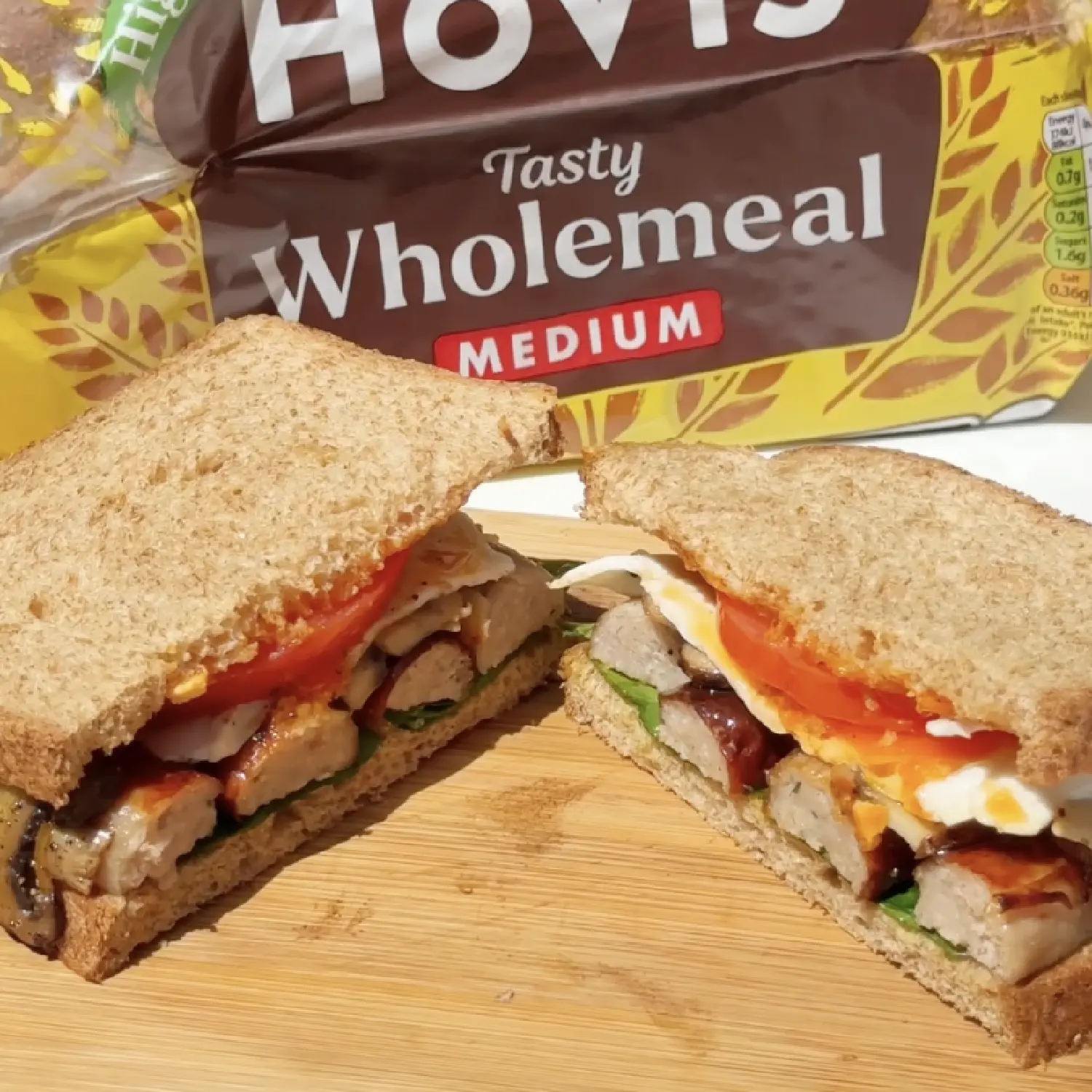 A sausage, egg and tomato sandwich cut in half on a wooden board in front of a loaf of Hovis Tasty Wholemeal bread.