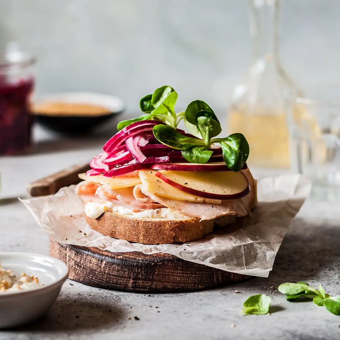 A delicious open-faced sandwich on a rustic wooden board, topped with sliced turkey or ham, cheese, thinly sliced red apple, and a vibrant pile of red onion and fresh greens. A small white bowl of sauce and a glass jar of what appears to be cranberry sauce are visible in the background, along with a bottle of oil or vinegar and a drinking glass. The setting appears to be a light and airy kitchen or dining area.