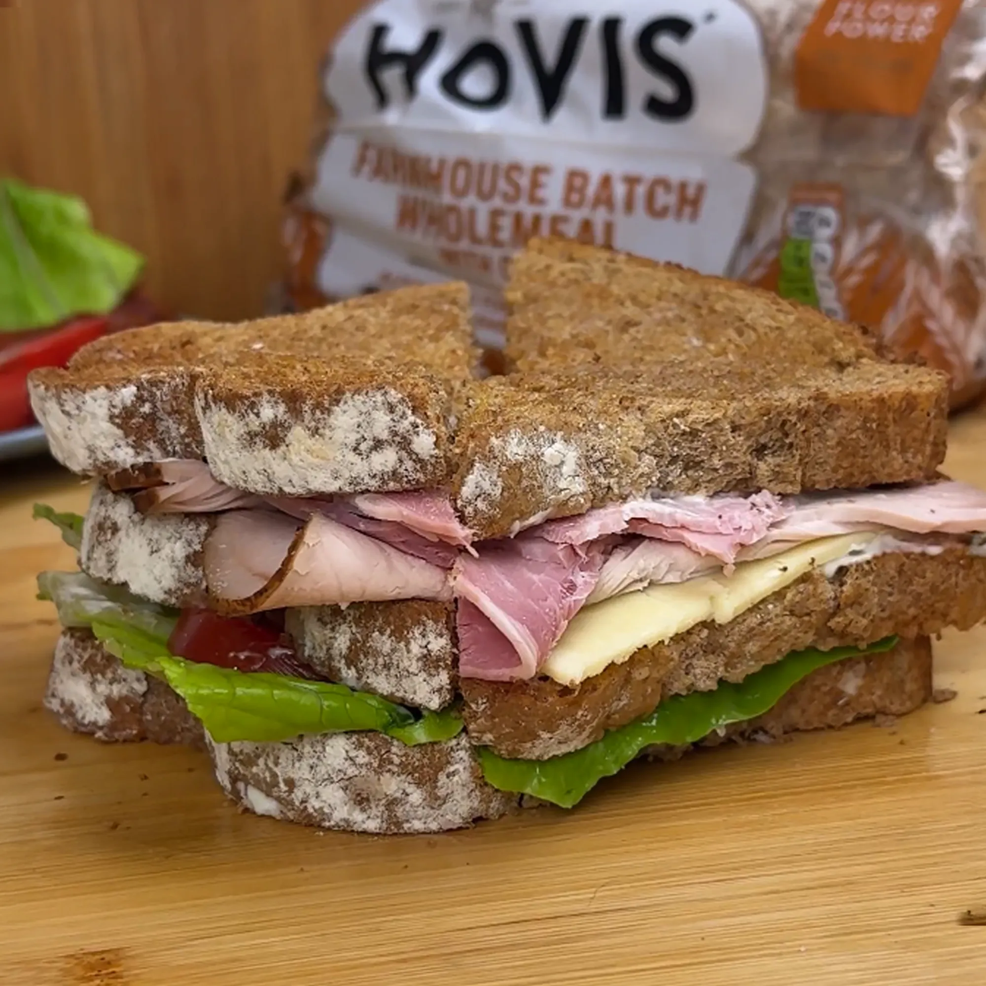 An image of the final Club sandwich with its ingredients spilling out, cut into half triangles on a cutting board. We see the Hovis® Farmhouse Batch Wholemeal loaf in the background.