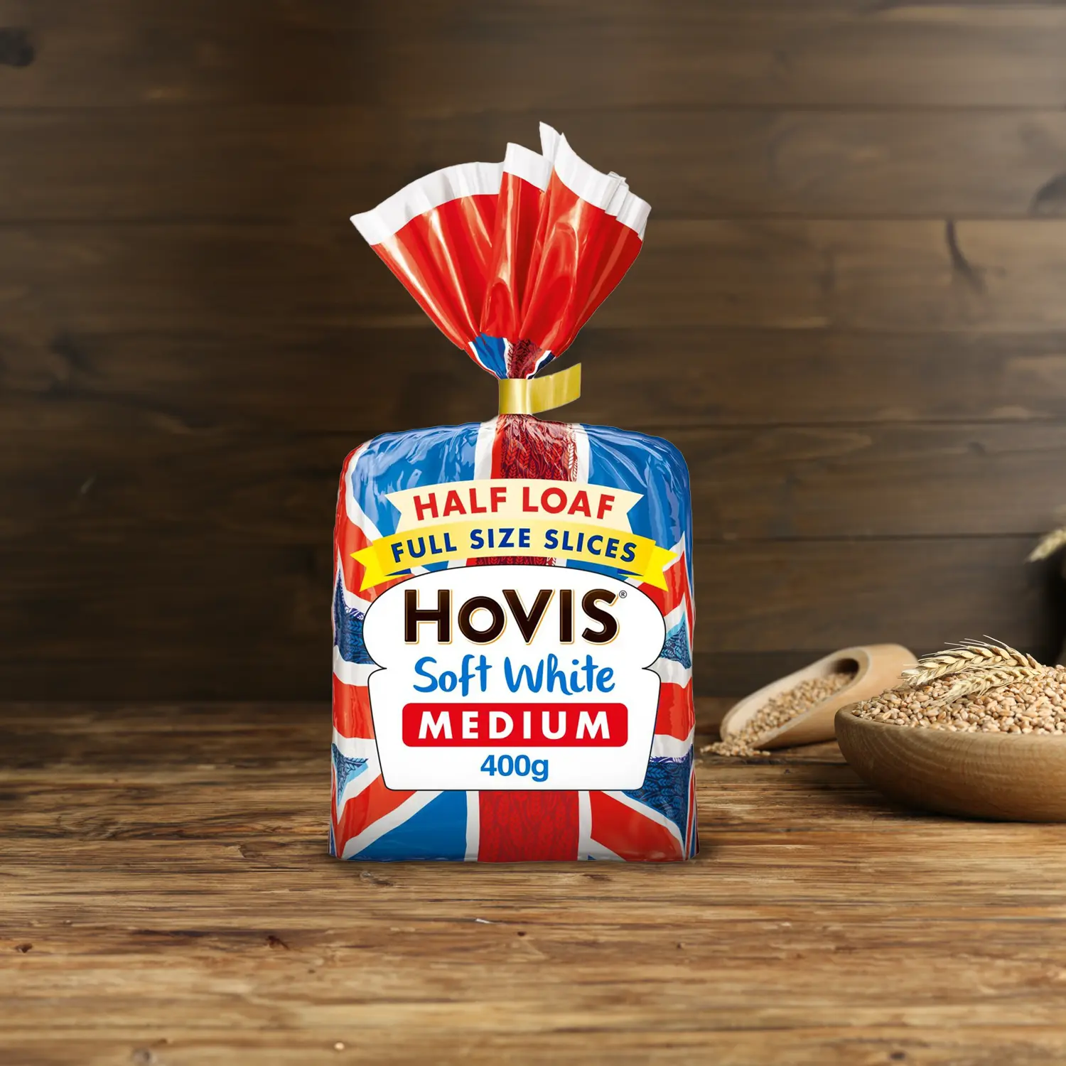 Hovis Soft White Medium 400g on a wooden table, with a bowl of wheat grains, a wooden scoop, and dried wheat in the background.