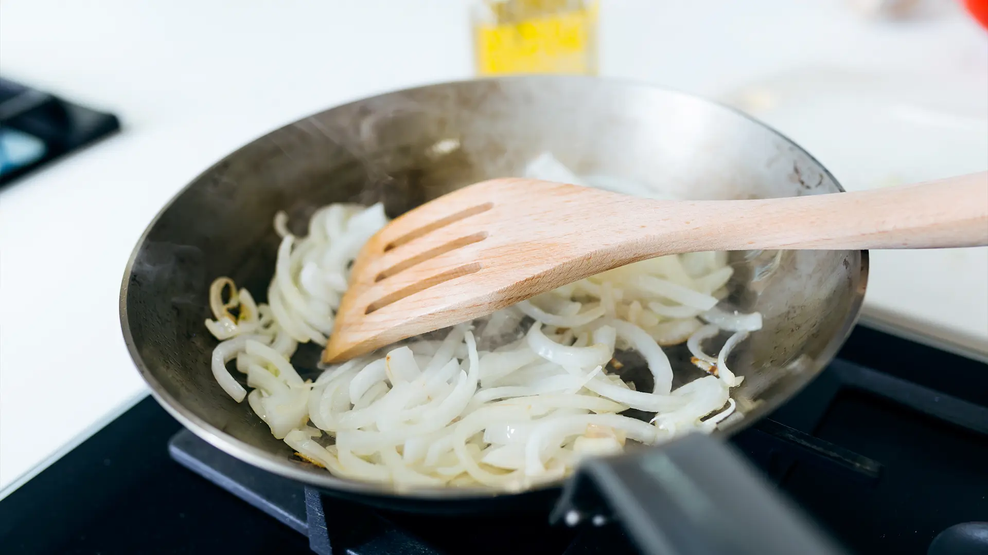 A close-up image of someone using a wooden spoon to stir sliced onions in a medium frying pan, slowly caramelising them.